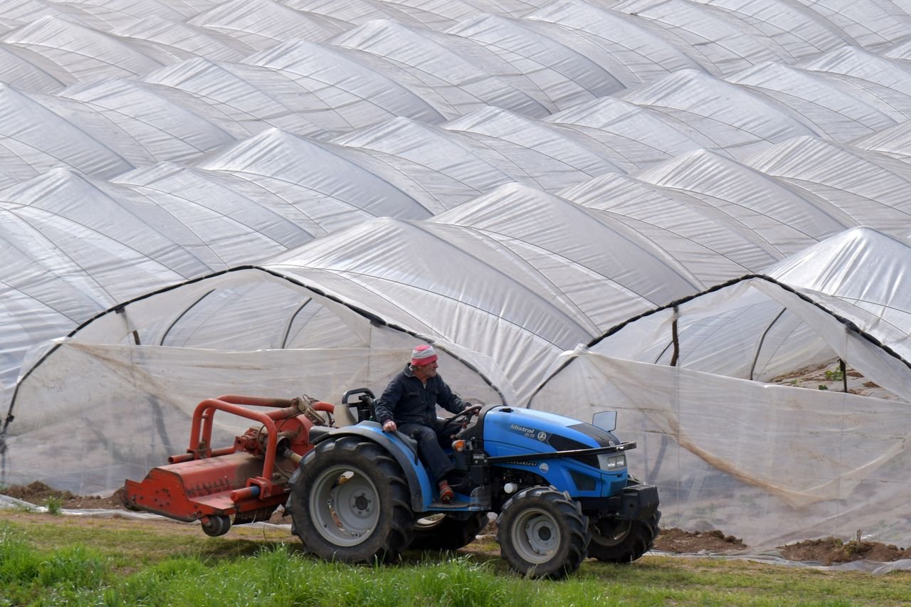 A farmer operates a blue tractor by greenhouses, showcasing modern agriculture.
